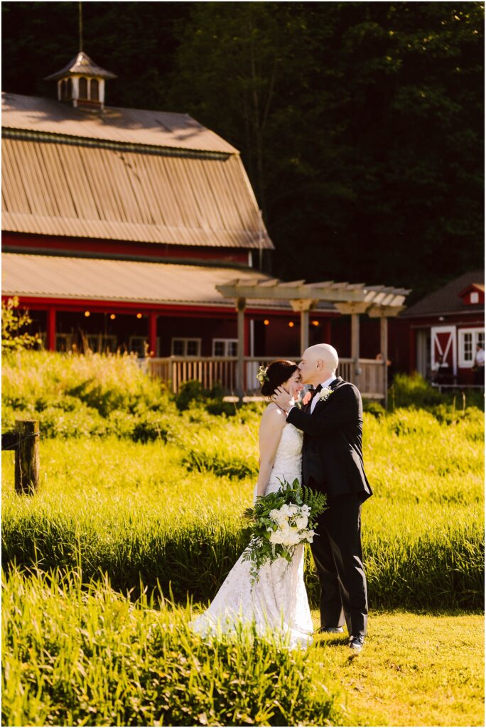 Snohomish Wedding Photographer GSquared Weddings A bride and groom stand in front of a rustic barn surrounded by lush greenery. The bride wears a lace gown and holds a bouquet, while the groom is in a dark suit. They gaze at each other lovingly under the warm sunlight. Serving Seattle, Snohomish and Washington State
