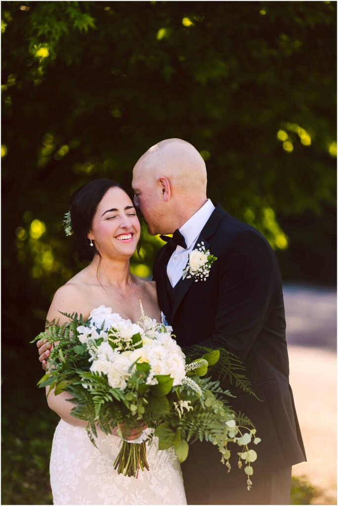 Snohomish Wedding Photographer GSquared Weddings A bride and groom stand outdoors, smiling. The groom, in a black tuxedo, leans in to whisper to the bride, who is wearing a white strapless gown and holding a bouquet of white flowers and greenery. They are surrounded by lush greenery. Serving Seattle, Snohomish and Washington State