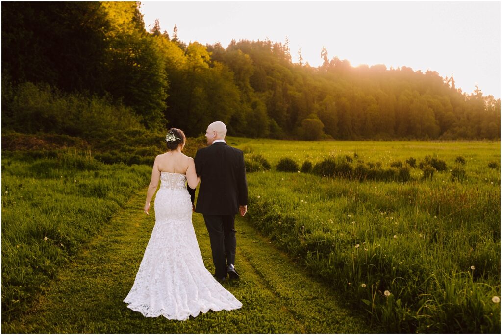 Snohomish Wedding Photographer GSquared Weddings A newlywed couple walks hand-in-hand down a grassy path, surrounded by a green meadow. The bride wears a white dress, and the groom is in a black suit. The sun sets behind trees in the background, casting a warm glow over the scene. Serving Seattle, Snohomish and Washington State