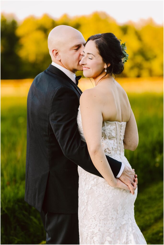 Snohomish Wedding Photographer GSquared Weddings A couple embraces in a grassy field, illuminated by warm sunlight. The man, wearing a black suit, gently kisses the womans face. She is in a white lace strapless dress, smiling with closed eyes. Trees are blurred in the background. Serving Seattle, Snohomish and Washington State