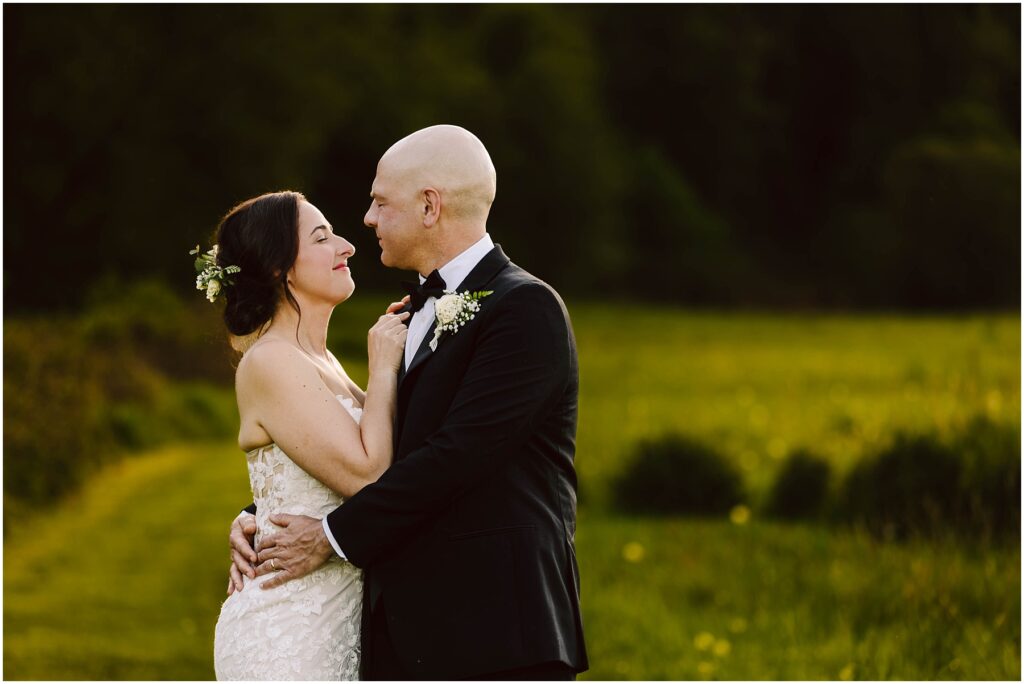 Snohomish Wedding Photographer GSquared Weddings A bride and groom embrace in a field with lush greenery in the background. The bride, in a white lace dress, smiles at the groom, who wears a black suit and bow tie. They are surrounded by a soft, sunlit landscape. Serving Seattle, Snohomish and Washington State