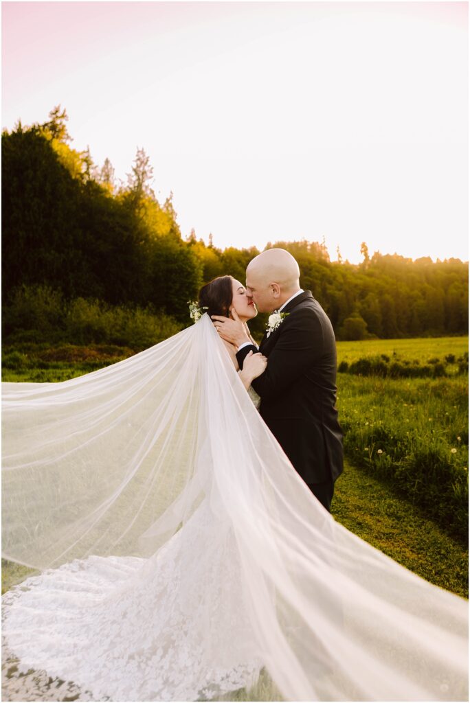Snohomish Wedding Photographer GSquared Weddings A bride and groom share a kiss in a scenic outdoor setting at sunset. The brides long veil flows dramatically in the breeze, creating a sense of movement. They are standing in a lush green field with trees in the background under a clear sky. Serving Seattle, Snohomish and Washington State
