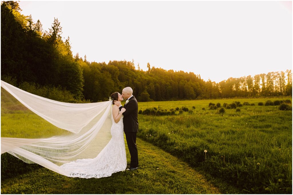 Snohomish Wedding Photographer GSquared Weddings A bride and groom stand in a grassy field at sunset, surrounded by trees. The bride wears a long, flowing veil, and the couple embraces, facing each other, with golden sunlight illuminating the scene. Serving Seattle, Snohomish and Washington State