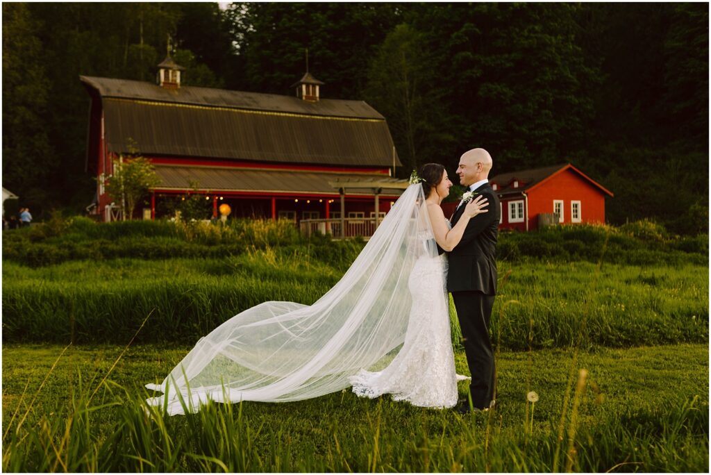 Snohomish Wedding Photographer GSquared Weddings Bride and groom embracing in a grassy field with a red barn in the background. The bride wears a long white gown with a flowing veil, while the groom is in a black suit. The barn has warm lights and a small red building nearby. Serving Seattle, Snohomish and Washington State