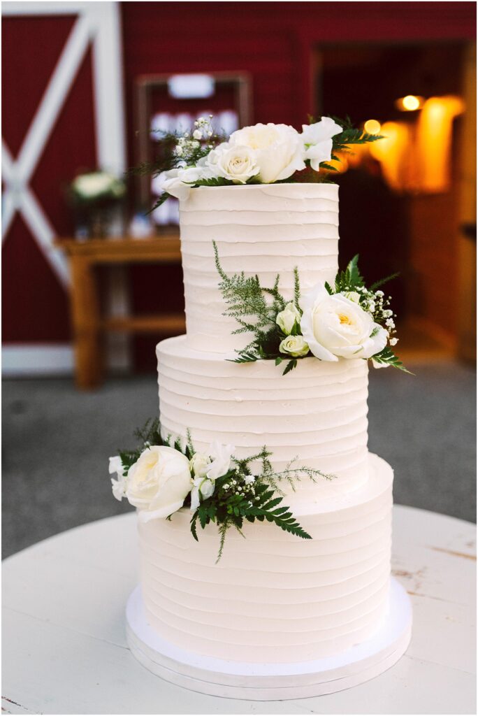 Snohomish Wedding Photographer GSquared Weddings Three-tiered white wedding cake adorned with white roses and green foliage. The cake features a ribbed texture and is displayed indoors, with a red and white wooden wall in the background. Serving Seattle, Snohomish and Washington State
