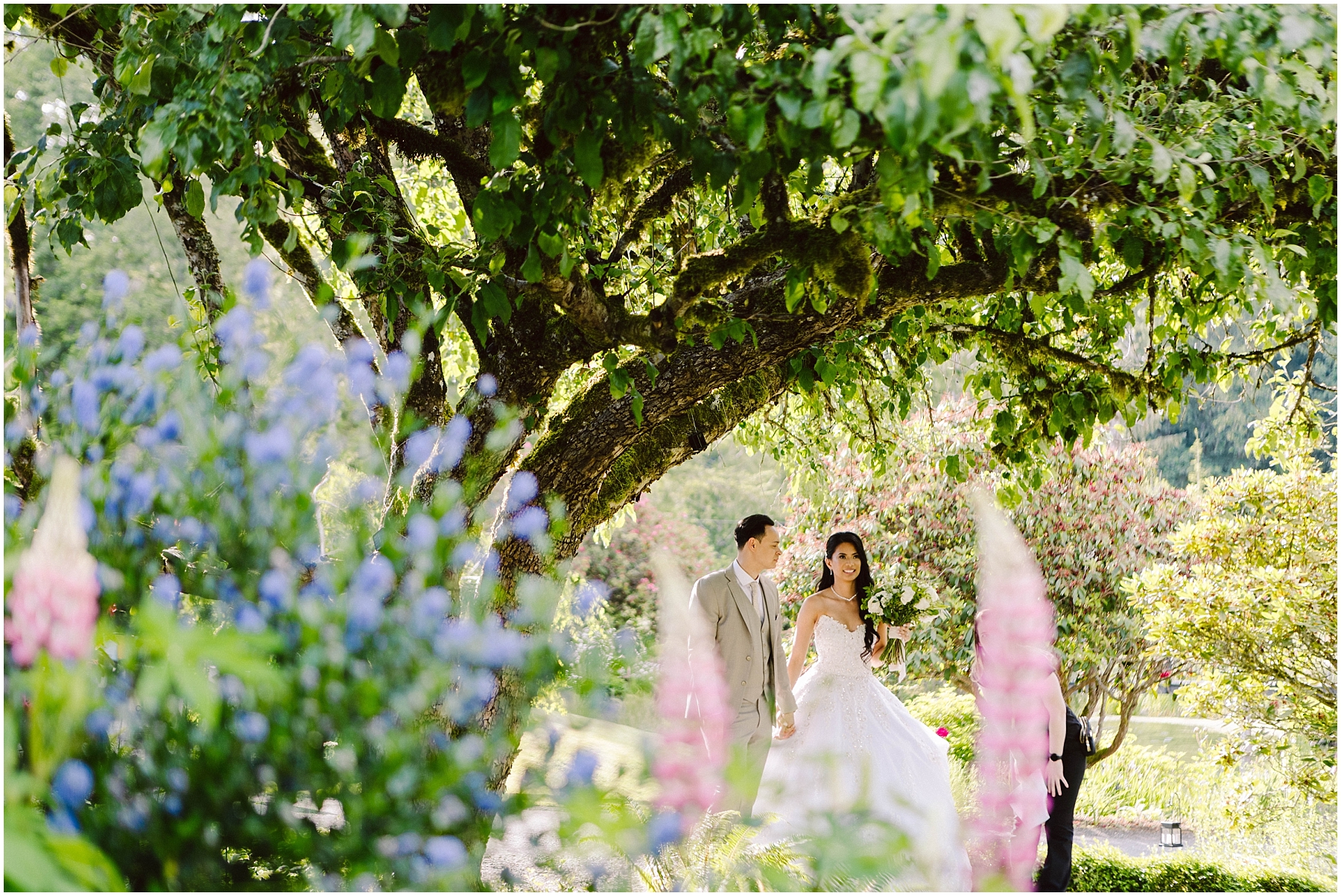 Snohomish Wedding Photographer GSquared Weddings A bride in a white wedding dress and a groom in a light-colored suit walk hand in hand under a large tree. They are surrounded by vibrant flowers and lush greenery, creating a romantic and picturesque garden setting. Serving Seattle, Snohomish and Washington State