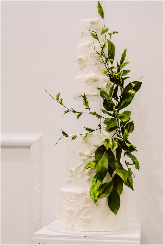 Snohomish Wedding Photographer GSquared Weddings A three-tiered white wedding cake decorated with textured floral patterns and adorned with cascading green leaves, displayed on a white pedestal against a plain background. Serving Seattle, Snohomish and Washington State