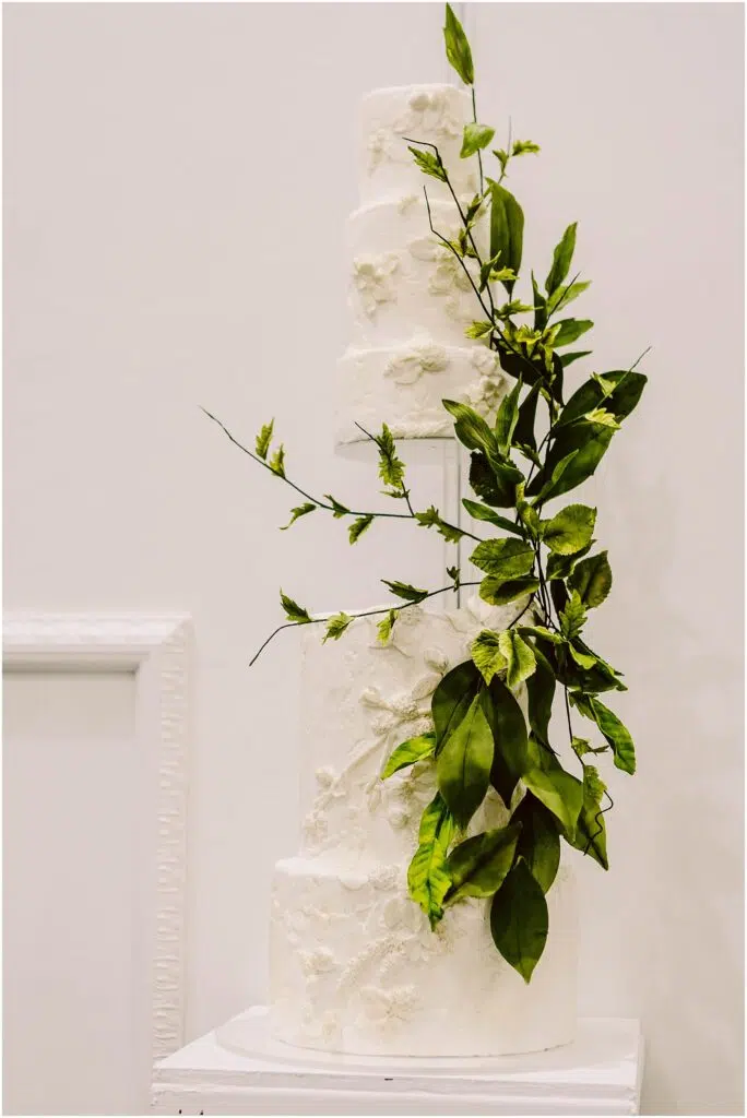 Snohomish Wedding Photographer GSquared Weddings A threetiered white wedding cake decorated with textured floral patterns and adorned with cascading green leaves displayed on a white pedestal against a plain background Serving Seattle Snohomish and Washington State Snohomish Wedding Photographer GSquared Weddings A three-tiered white wedding cake decorated with textured floral patterns and adorned with cascading green leaves, displayed on a white pedestal against a plain background. Serving Seattle, Snohomish and Washington State