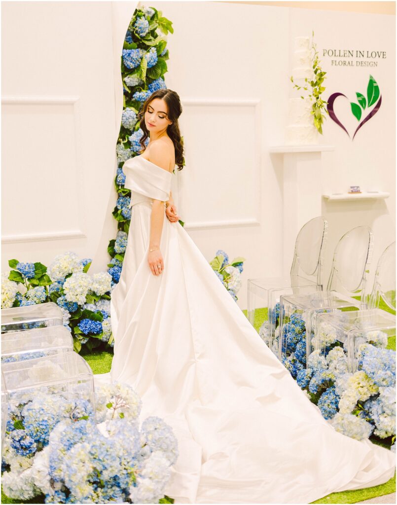 Snohomish Wedding Photographer GSquared Weddings A woman in an elegant off-the-shoulder white gown poses beside a display of blue and white floral arrangements. The setting includes clear chairs and a sign that reads Pollen in Love Floral Design with a heart-shaped logo. Serving Seattle, Snohomish and Washington State