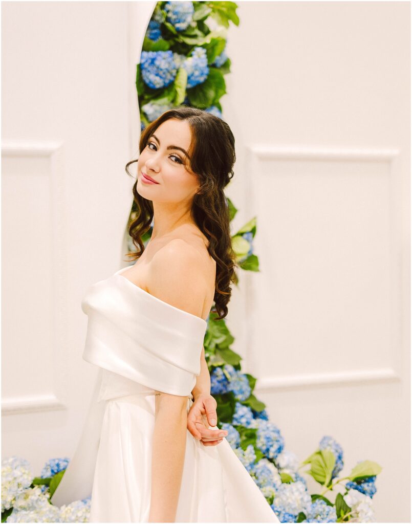 Snohomish Wedding Photographer GSquared Weddings A woman in an elegant off-shoulder white dress stands gracefully in front of a backdrop decorated with blue and white hydrangeas. She has long, wavy brown hair and is smiling softly, creating a serene and joyful atmosphere. Serving Seattle, Snohomish and Washington State