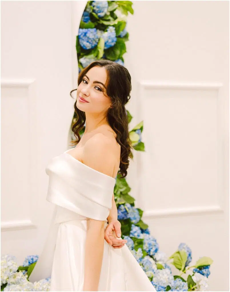 Snohomish Wedding Photographer GSquared Weddings A woman in an elegant offshoulder white dress stands gracefully in front of a backdrop decorated with blue and white hydrangeas She has long wavy brown hair and is smiling softly creating a serene and joyful atmosphere Serving Seattle Snohomish and Washington State Snohomish Wedding Photographer GSquared Weddings A woman in an elegant off-shoulder white dress stands gracefully in front of a backdrop decorated with blue and white hydrangeas. She has long, wavy brown hair and is smiling softly, creating a serene and joyful atmosphere. Serving Seattle, Snohomish and Washington State
