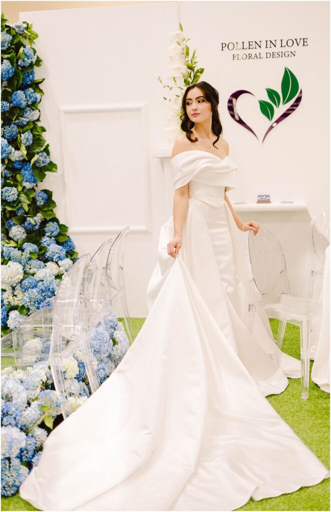 Snohomish Wedding Photographer GSquared Weddings Bride in an elegant white gown with a long train stands in front of a floral display featuring blue and white flowers. Clear chairs line the area. A heart-shaped floral design and the words Pollen in Love Floral Design are on the wall. Serving Seattle, Snohomish and Washington State