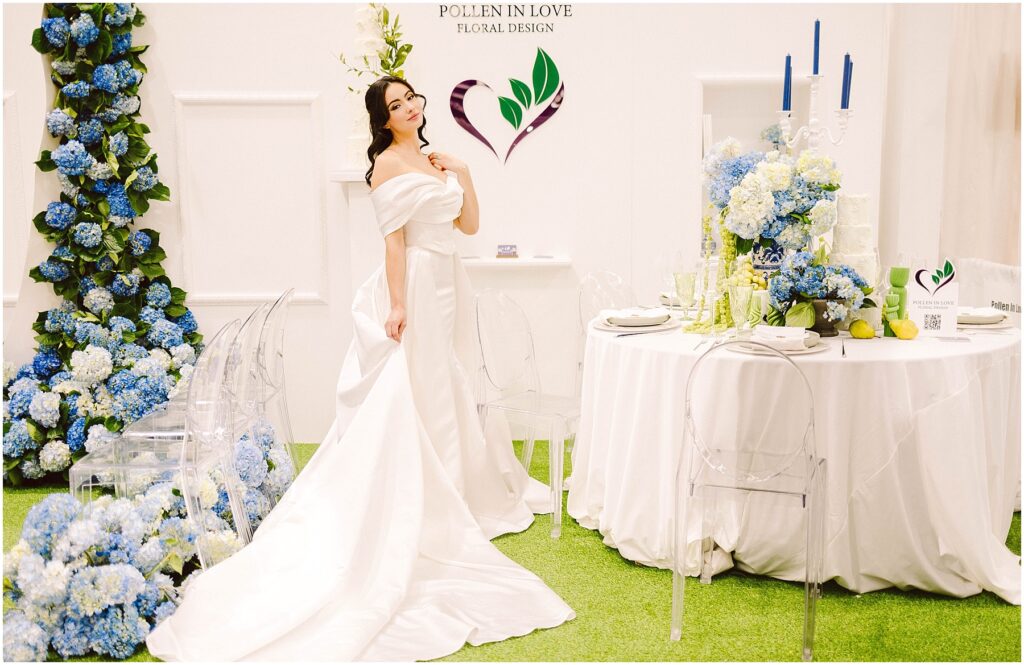 Snohomish Wedding Photographer GSquared Weddings A woman in an elegant white gown stands in a floral-themed setup, next to a white round table with blue floral and candle arrangements. Pollen in Love Floral Design is displayed on the wall. Clear chairs surround the table on a grass-like carpet. Serving Seattle, Snohomish and Washington State