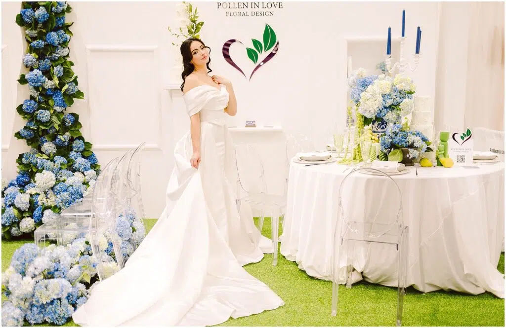 Snohomish Wedding Photographer GSquared Weddings A woman in an elegant white gown stands in a floralthemed setup next to a white round table with blue floral and candle arrangements Pollen in Love Floral Design is displayed on the wall Clear chairs surround the table on a grasslike carpet Serving Seattle Snohomish and Washington State Snohomish Wedding Photographer GSquared Weddings A woman in an elegant white gown stands in a floral-themed setup, next to a white round table with blue floral and candle arrangements. Pollen in Love Floral Design is displayed on the wall. Clear chairs surround the table on a grass-like carpet. Serving Seattle, Snohomish and Washington State