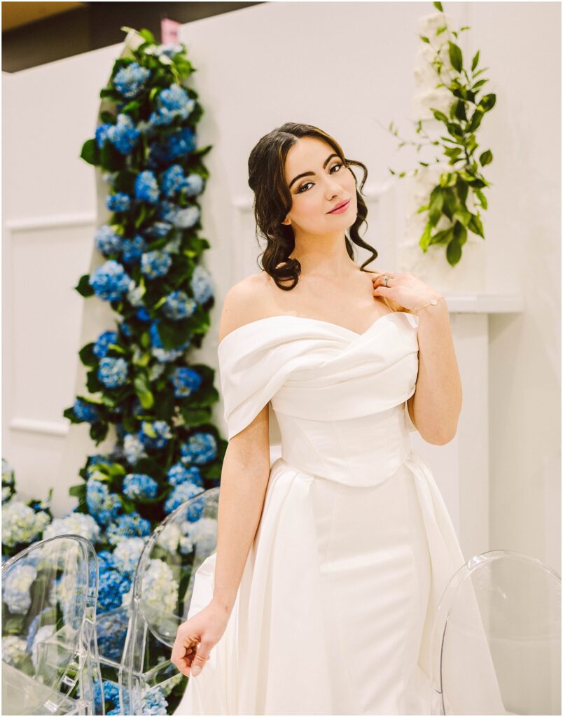 Snohomish Wedding Photographer GSquared Weddings A woman in an elegant off-shoulder white dress stands gracefully beside a tall floral arrangement of blue and white flowers. She is in a well-lit setting with clear chairs visible nearby, exuding a serene and poised demeanor. Serving Seattle, Snohomish and Washington State
