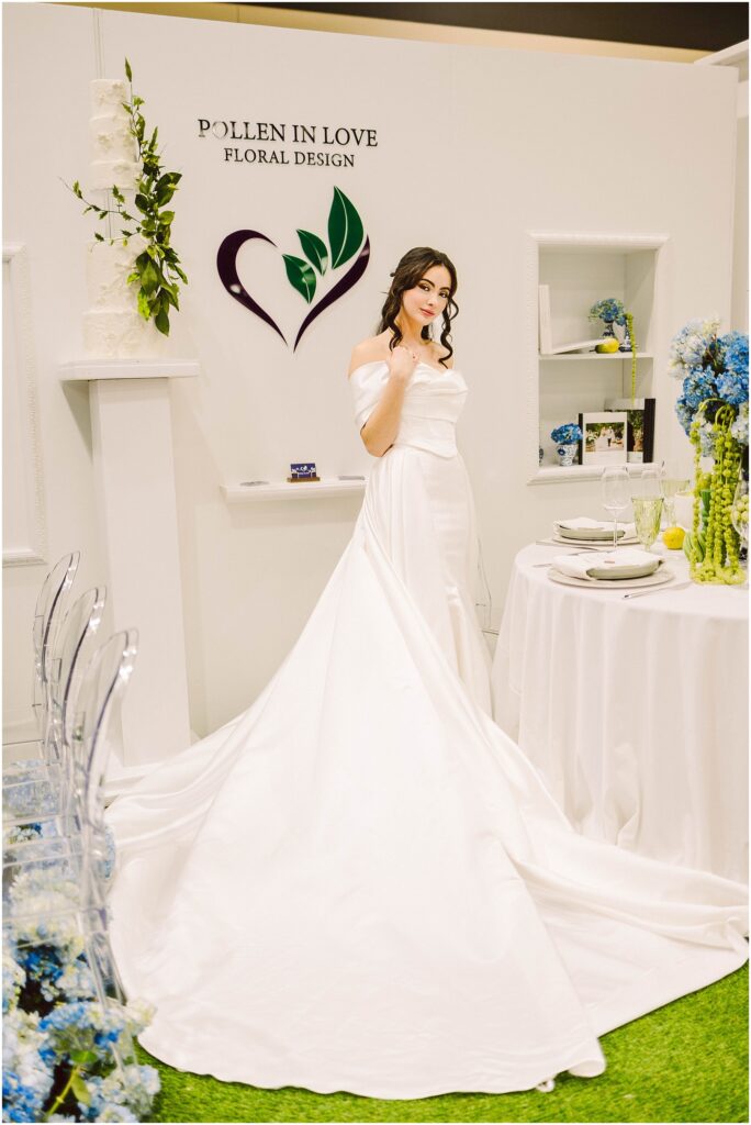 Snohomish Wedding Photographer GSquared Weddings A woman in a white wedding dress poses in front of a Pollen in Love Floral Design sign. The setting includes a table with blue flowers, elegant tableware, and a long train extending from the dress. The space has a sophisticated floral theme. Serving Seattle, Snohomish and Washington State
