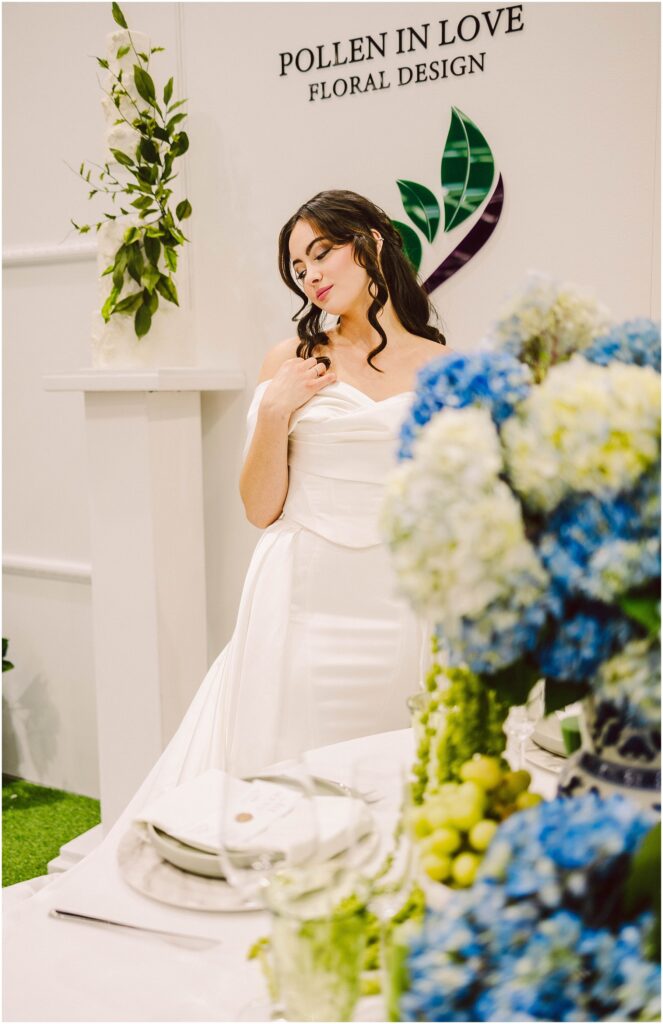 Snohomish Wedding Photographer GSquared Weddings A woman in an elegant white dress stands beside a table with blue and green floral arrangements. She looks down thoughtfully, with her hand near her shoulder. The backdrop features the words Pollen in Love Floral Design and leafy decorations. Serving Seattle, Snohomish and Washington State
