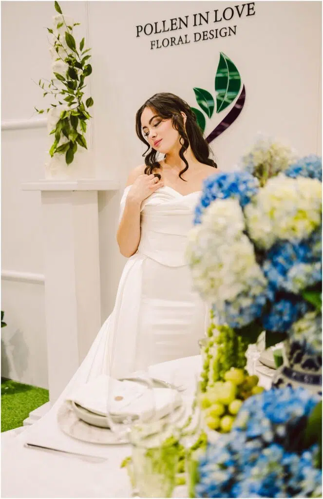 Snohomish Wedding Photographer GSquared Weddings A woman in an elegant white dress stands beside a table with blue and green floral arrangements She looks down thoughtfully with her hand near her shoulder The backdrop features the words Pollen in Love Floral Design and leafy decorations Serving Seattle Snohomish and Washington State Snohomish Wedding Photographer GSquared Weddings A woman in an elegant white dress stands beside a table with blue and green floral arrangements. She looks down thoughtfully, with her hand near her shoulder. The backdrop features the words Pollen in Love Floral Design and leafy decorations. Serving Seattle, Snohomish and Washington State