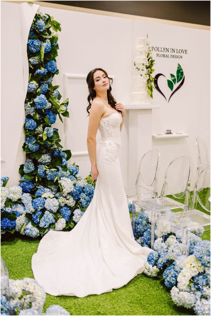 Snohomish Wedding Photographer GSquared Weddings A bride in a strapless white wedding dress poses gracefully beside floral arrangements of blue and white hydrangeas. Clear chairs and a floral design logo are in the background. Serving Seattle, Snohomish and Washington State