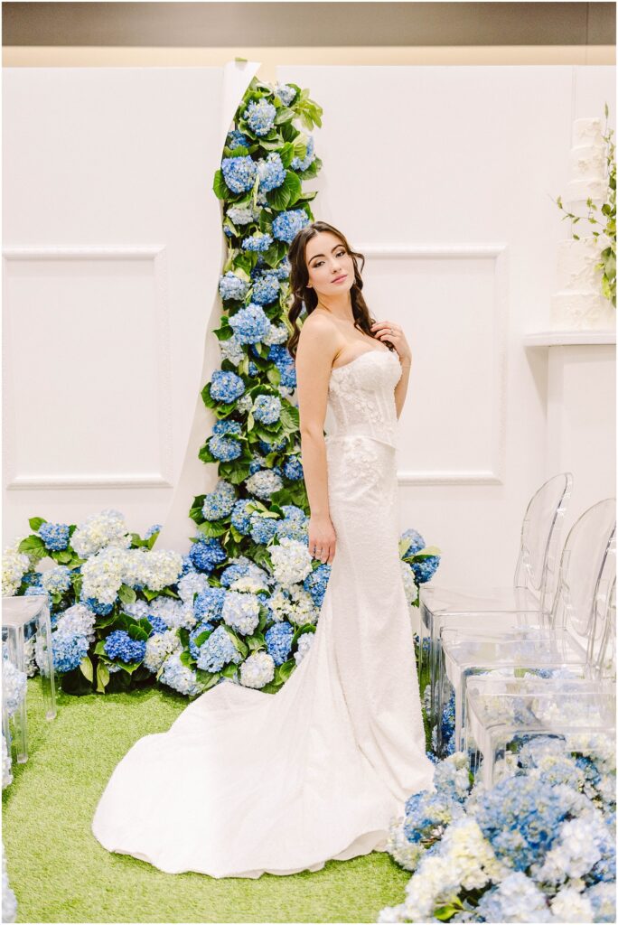 Snohomish Wedding Photographer GSquared Weddings A bride in a strapless white gown stands by a floral arch adorned with blue and white hydrangeas. She poses on green grass next to clear chairs, with her hair styled in loose waves. The setting is elegant and bright. Serving Seattle, Snohomish and Washington State