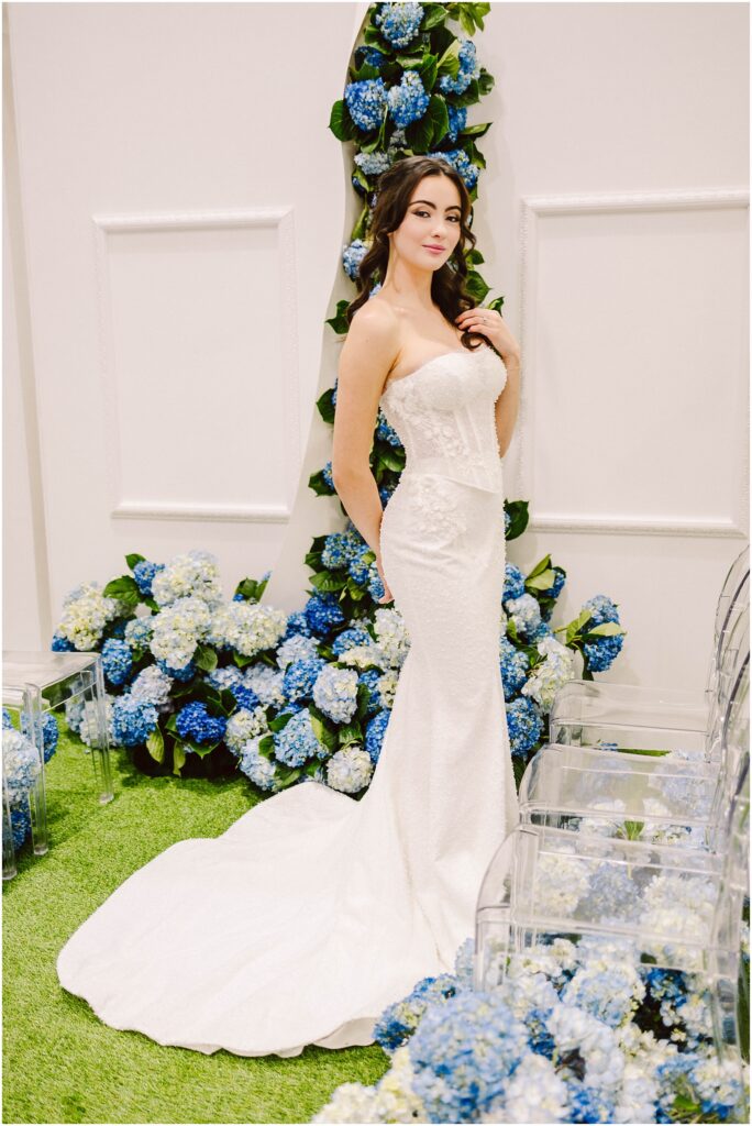 Snohomish Wedding Photographer GSquared Weddings A woman in a strapless white wedding dress stands on green grass. She is surrounded by blue and white hydrangea flowers. Clear chairs are visible on the side. The setting is elegant and floral. Serving Seattle, Snohomish and Washington State