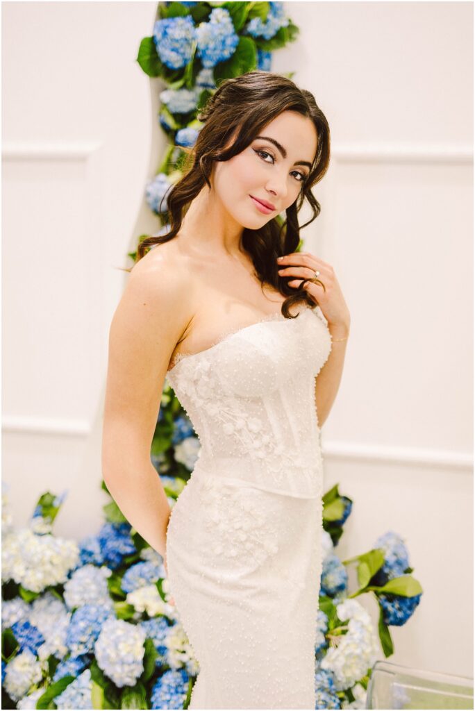 Snohomish Wedding Photographer GSquared Weddings A woman in a strapless white wedding dress stands in front of a backdrop adorned with blue and white flowers. She has long, wavy hair and is posing with one hand on her hip, smiling softly at the camera. Serving Seattle, Snohomish and Washington State