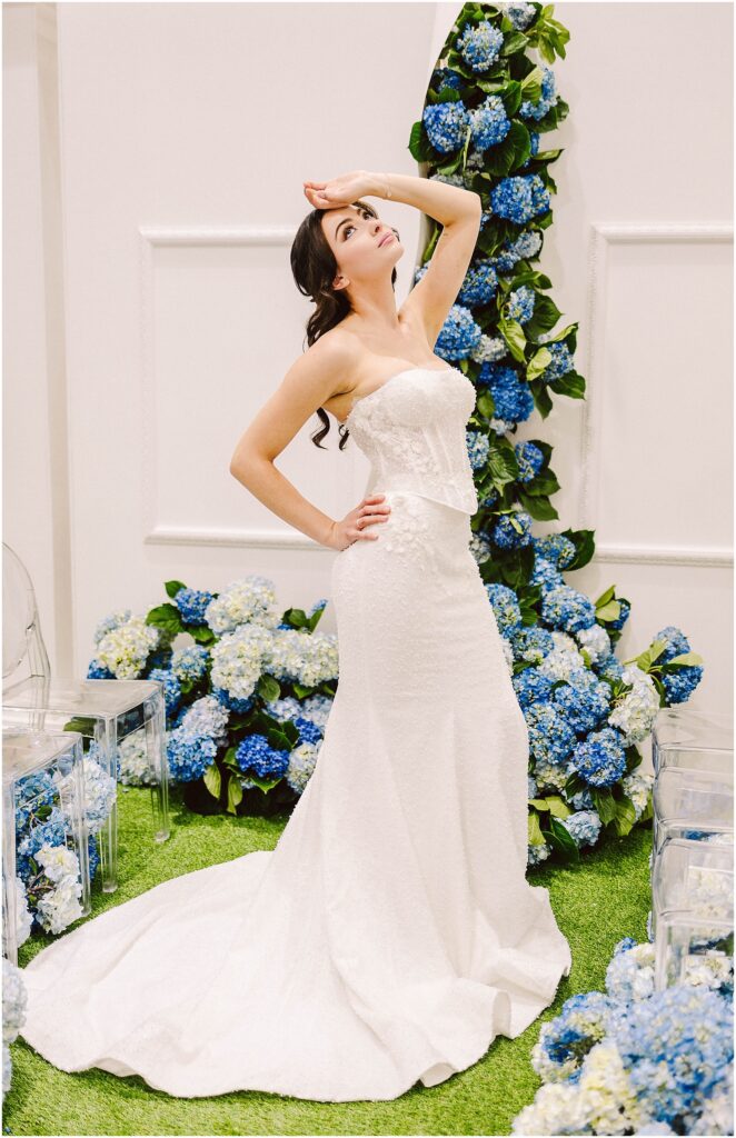 Snohomish Wedding Photographer GSquared Weddings A woman in a strapless white wedding dress poses with one hand on her hip and the other on her forehead. She stands on grass surrounded by blue and white flowers. Transparent chairs are visible nearby. Serving Seattle, Snohomish and Washington State