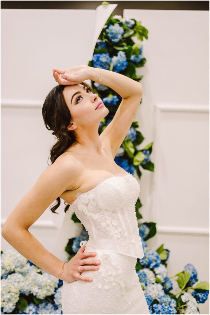 Snohomish Wedding Photographer GSquared Weddings A bride in a strapless, white lace wedding dress poses dramatically. Her hand rests on her forehead, and she stands in front of a backdrop adorned with blue and white flowers. Her dark hair is styled elegantly. Serving Seattle, Snohomish and Washington State