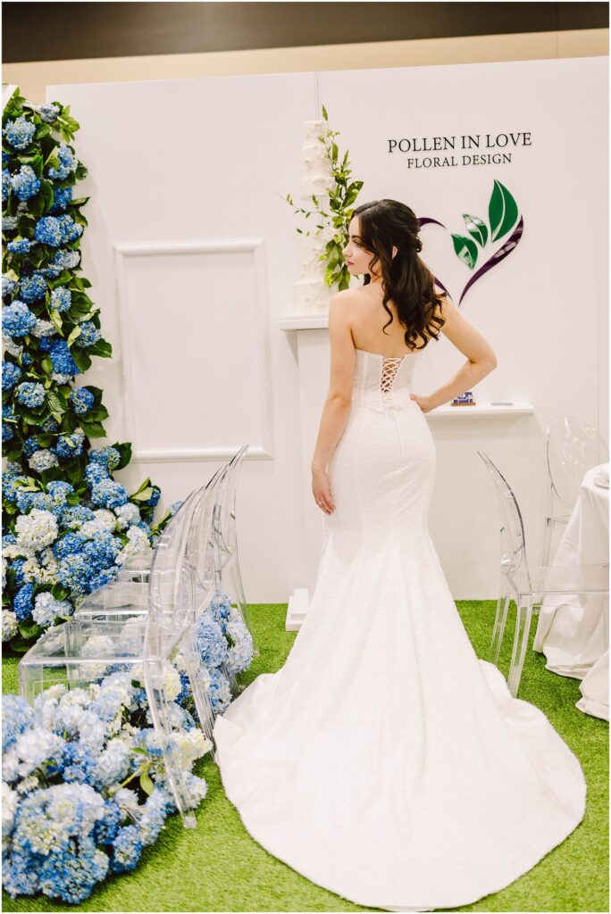 Snohomish Wedding Photographer GSquared Weddings A bride in a strapless white gown stands with her back to the camera, in front of a display with blue hydrangeas and a white floral-themed backdrop. The setting includes transparent chairs and a sign reading Pollen in Love Floral Design. Serving Seattle, Snohomish and Washington State