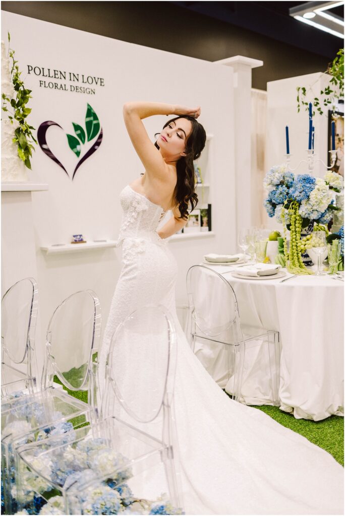Snohomish Wedding Photographer GSquared Weddings A woman in a strapless white wedding gown poses with her arm raised, standing next to a table with a white tablecloth and blue flowers. Transparent chairs surround the table. The backdrop features floral designs and the words Pollen In Love Floral Design. Serving Seattle, Snohomish and Washington State