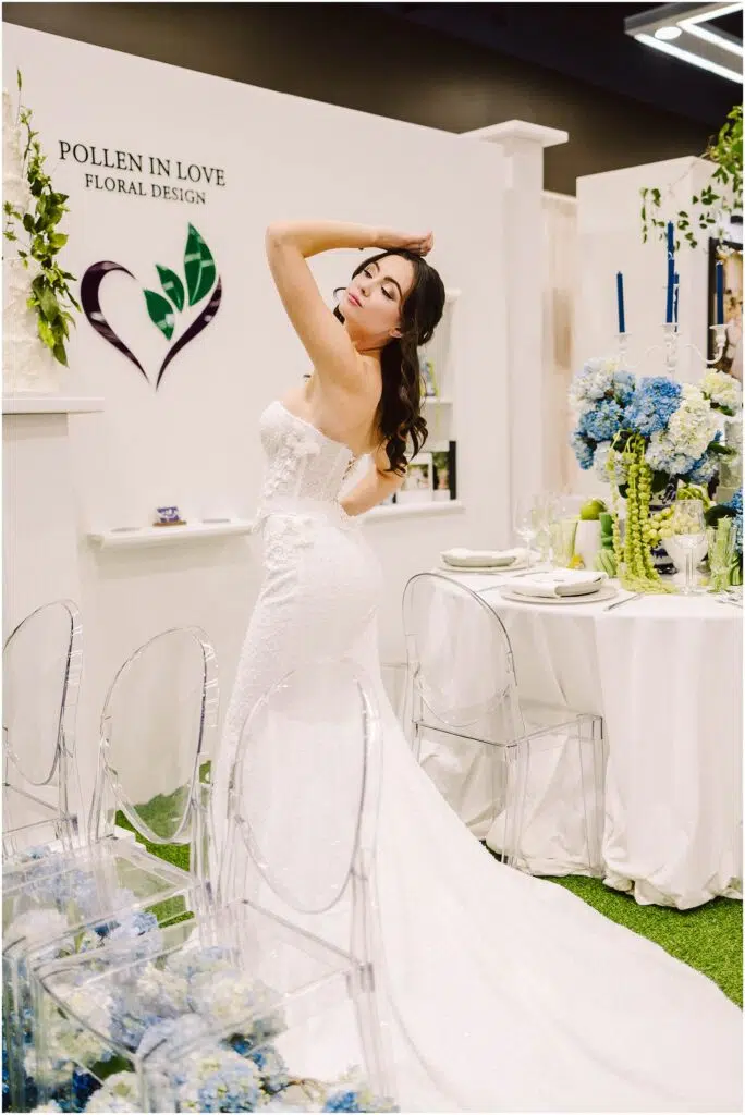 Snohomish Wedding Photographer GSquared Weddings A woman in a strapless white wedding gown poses with her arm raised standing next to a table with a white tablecloth and blue flowers Transparent chairs surround the table The backdrop features floral designs and the words Pollen In Love Floral Design Serving Seattle Snohomish and Washington State Snohomish Wedding Photographer GSquared Weddings A woman in a strapless white wedding gown poses with her arm raised, standing next to a table with a white tablecloth and blue flowers. Transparent chairs surround the table. The backdrop features floral designs and the words Pollen In Love Floral Design. Serving Seattle, Snohomish and Washington State