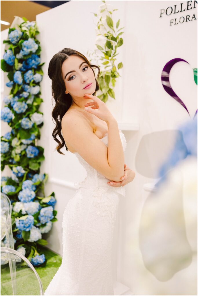 Snohomish Wedding Photographer GSquared Weddings A woman in a white strapless dress stands in a floral-themed setting with blue and green hydrangeas. She is gazing at the camera with one hand touching her face. The background features floral decor and a partial logo. Serving Seattle, Snohomish and Washington State