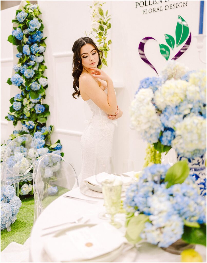 Snohomish Wedding Photographer GSquared Weddings A woman in a white dress poses elegantly beside a floral display of blue hydrangeas. She stands near a table set with blue and white flowers, clear chairs, and elegant tableware. A floral design logo is visible on the wall behind her. Serving Seattle, Snohomish and Washington State