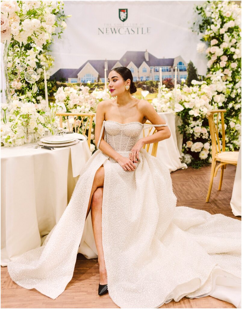 Snohomish Wedding Photographer GSquared Weddings A woman in an elegant strapless white gown with a long train sits gracefully at a decorated table. The background features lush floral arrangements and an image of a grand building. She wears her hair up and has a thoughtful expression. Serving Seattle, Snohomish and Washington State