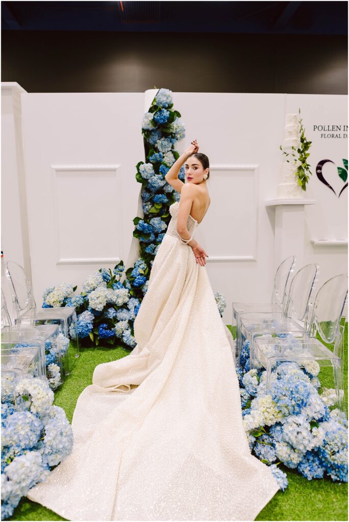 Snohomish Wedding Photographer GSquared Weddings Bride in a sparkling gown poses elegantly among clusters of blue and white hydrangeas. Clear chairs and white walls border the scene, creating a sophisticated wedding setting. Serving Seattle, Snohomish and Washington State