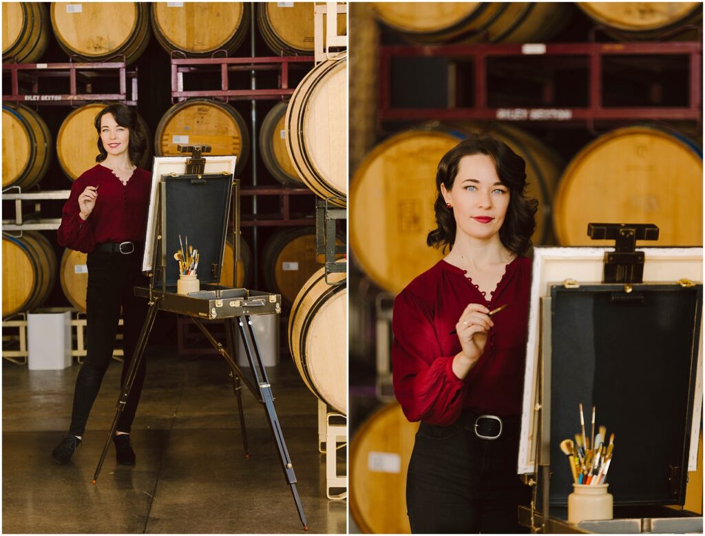 Snohomish Wedding Photographer GSquared Weddings A woman with dark hair, wearing a red blouse and black pants, stands by an easel in a winery or cellar. She holds a brush and poses confidently in front of wooden barrels. Brushes and paints are visible on the easel. Serving Seattle, Snohomish and Washington State