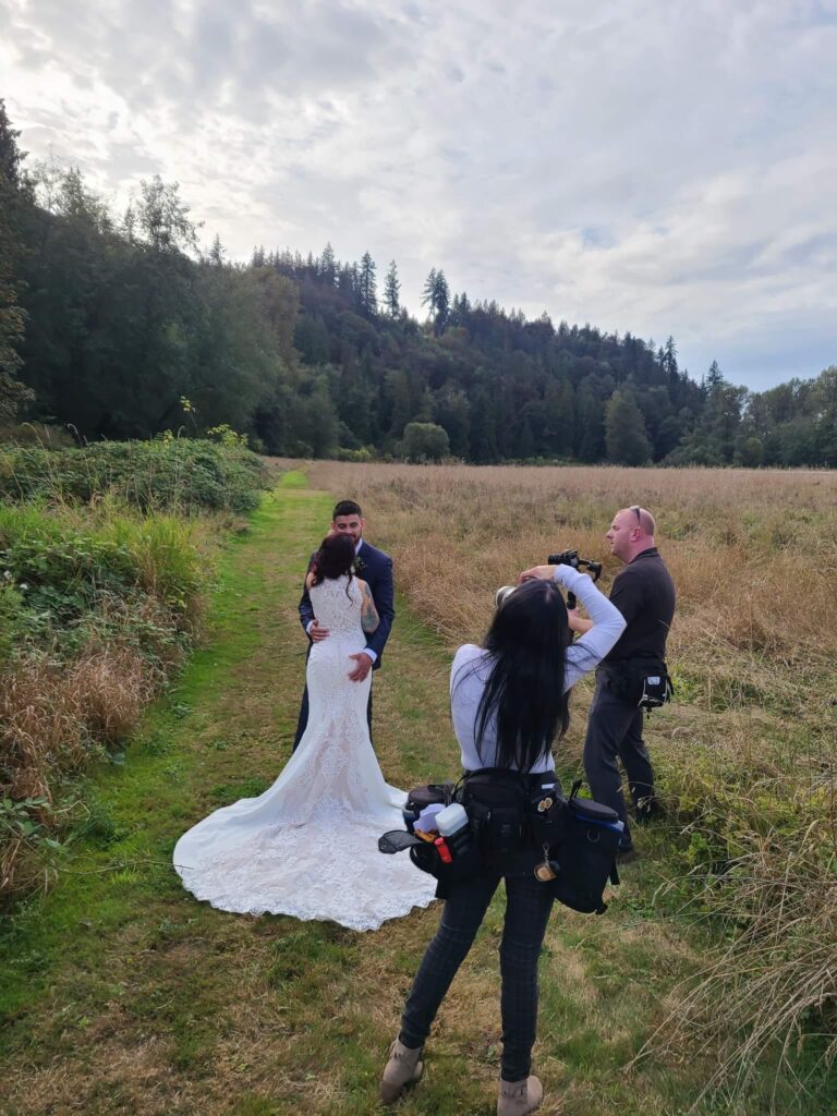 Snohomish Wedding Photographer GSquared Weddings A couple in wedding attire embraces on a grassy path surrounded by tall grass and trees. A photographer and assistant capture the moment. The sky is cloudy, and a forested hillside is in the background. Serving Seattle, Snohomish and Washington State