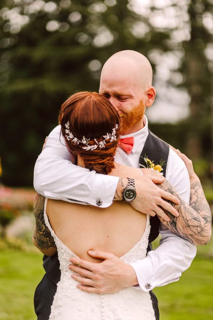 Snohomish Wedding Photographer GSquared Weddings A bald man in a vest tenderly embraces a woman in a white dress adorned with an intricate hair accessory. Outdoors, amidst lush greenery, he appears on the verge of tears, his eyes closed and face showing a deeply emotional expression. Serving Seattle, Snohomish and Washington State