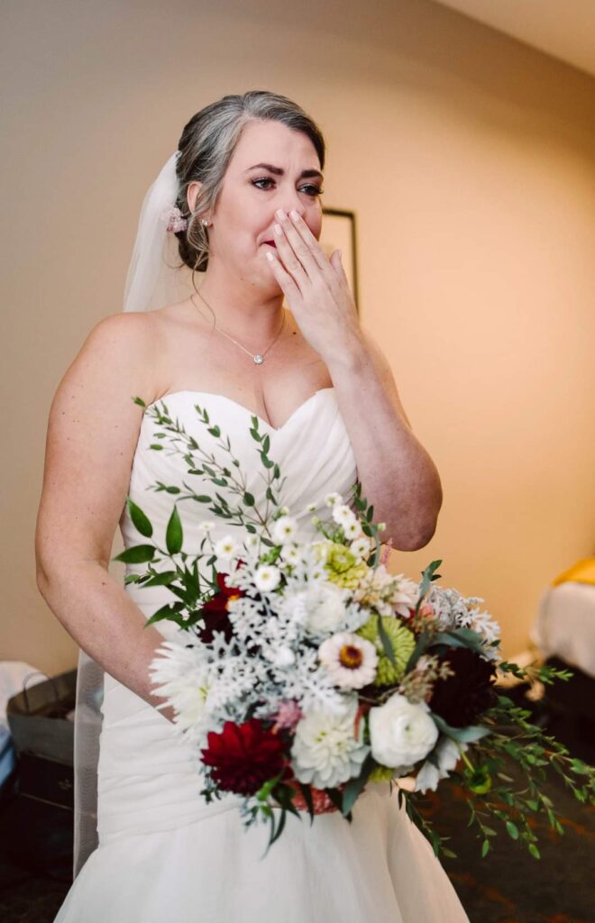 Snohomish Wedding Photographer GSquared Weddings A bride in a white dress and veil stands indoors, holding a bouquet of white, red, and green flowers. She appears emotional, on the verge of tears as she covers her mouth with one hand. Serving Seattle, Snohomish and Washington State