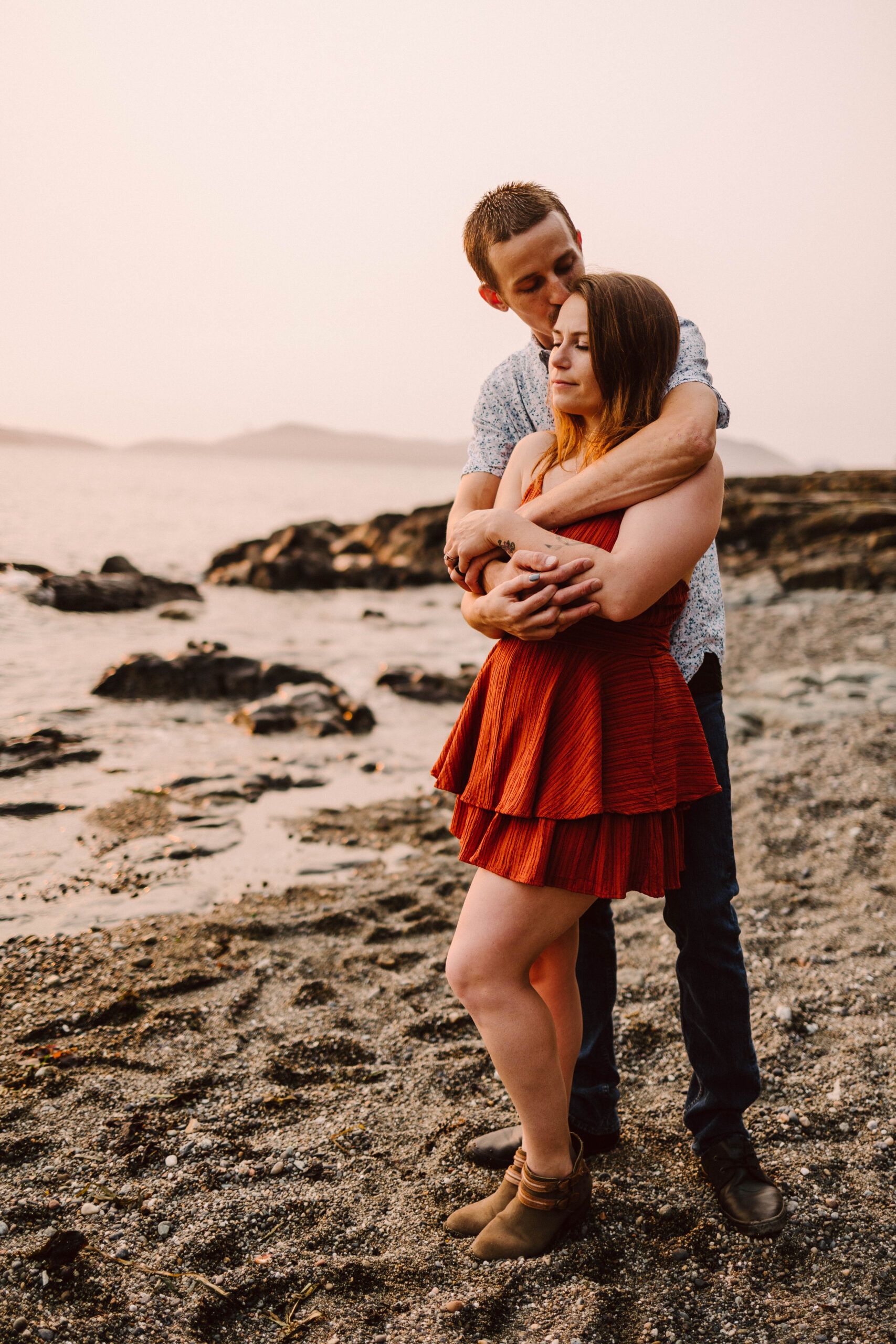 Snohomish Wedding Photographer GSquared Weddings A couple stands on a rocky beach at sunset capturing a perfect photo moment The man in a patterned shirt and jeans embraces the woman from behind She wears a red dress and boots eyes closed against the backdrop of the ocean and distant hills Serving Seattle Snohomish and Washington State