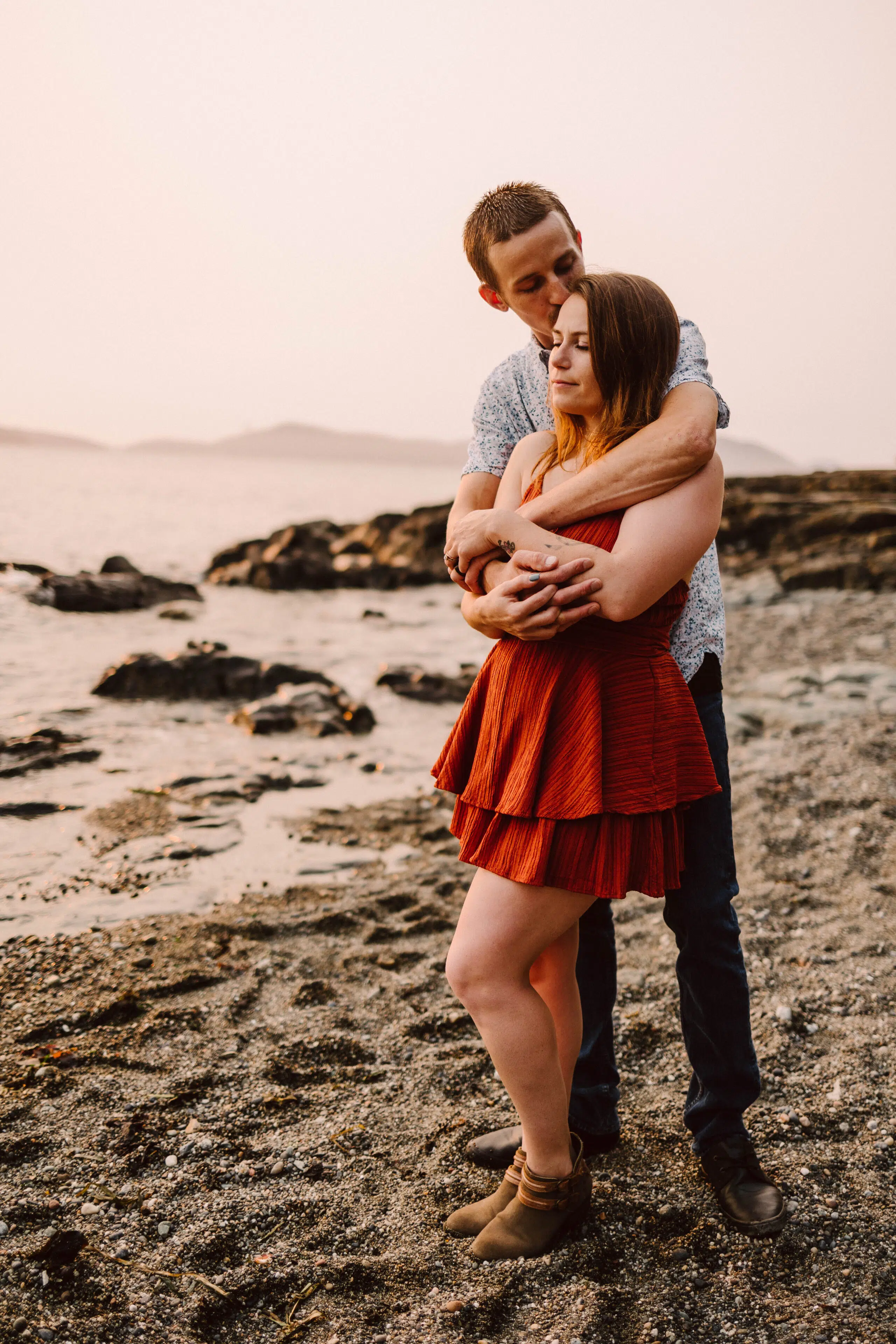 Snohomish Wedding Photographer GSquared Weddings A couple stands on a rocky beach at sunset capturing a perfect photo moment The man in a patterned shirt and jeans embraces the woman from behind She wears a red dress and boots eyes closed against the backdrop of the ocean and distant hills Serving Seattle Snohomish and Washington State