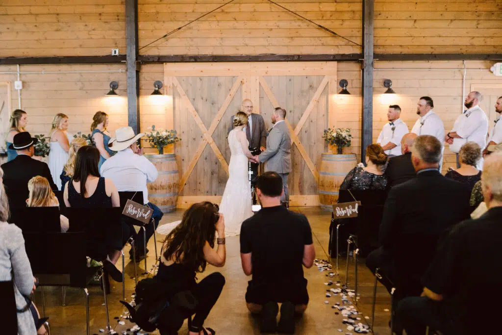 Snohomish Wedding Photographer GSquared Weddings A couple stands at the altar in a rustic setting exchanging vows surrounded by their wedding party and guests The venue features wooden walls and decor with a photographer capturing the moment Serving Seattle Snohomish and Washington State