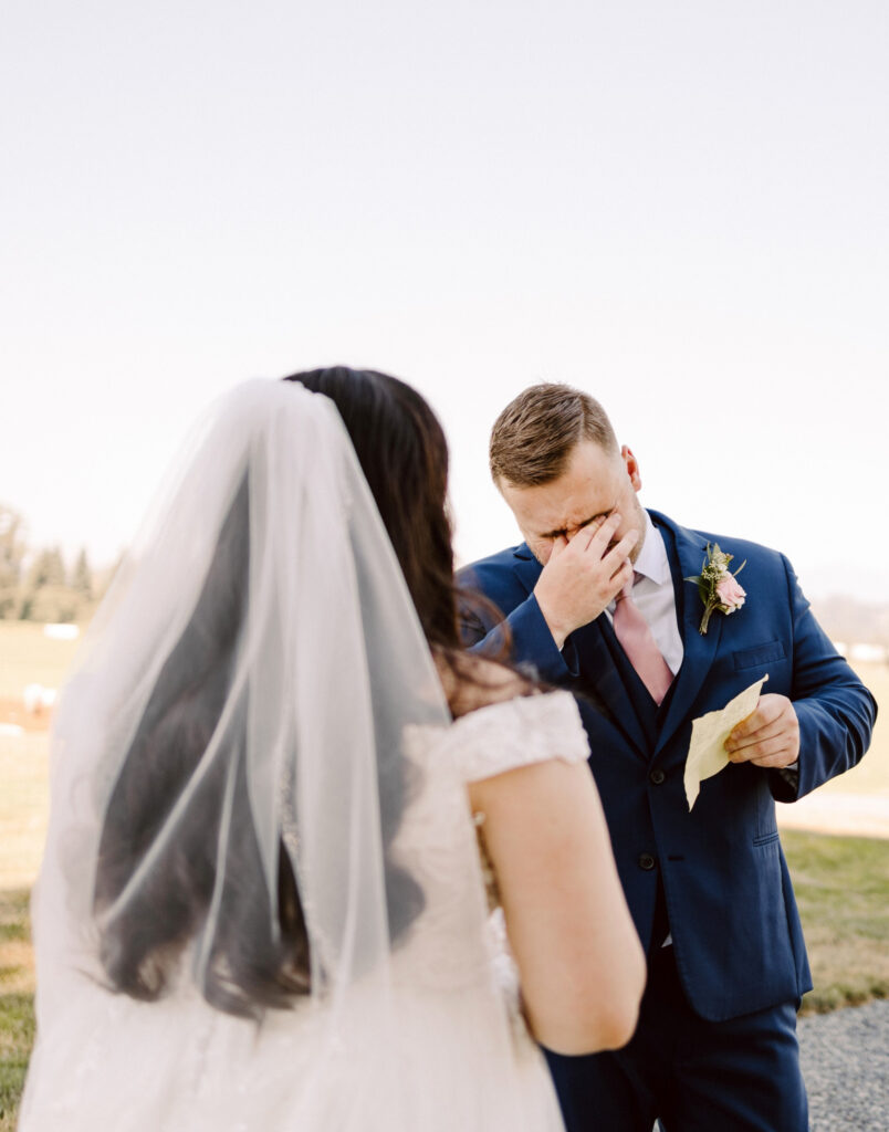 Snohomish Wedding Photographer GSquared Weddings A groom in a blue suit wipes his teary eyes while holding a handwritten note, standing outdoors. A bride, with long hair and a veil, faces him in her white dress. The background reveals a grassy field under a clear sky. Serving Seattle, Snohomish and Washington State