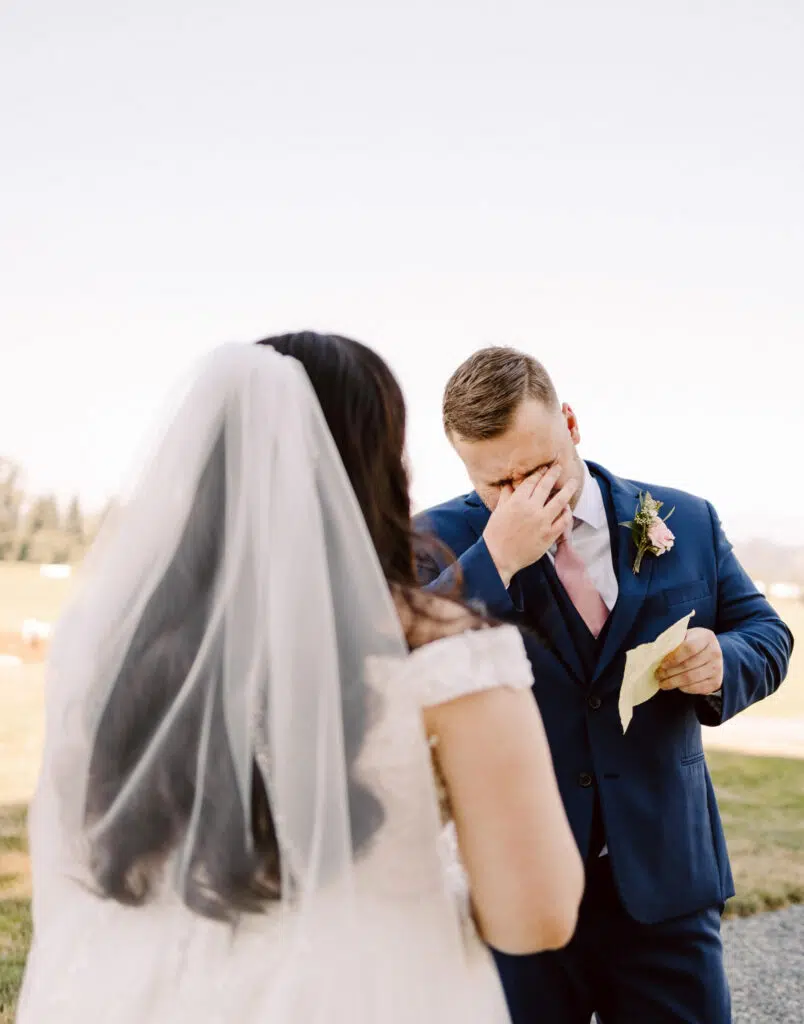 Snohomish Wedding Photographer GSquared Weddings A groom in a blue suit wipes his teary eyes while holding a handwritten note standing outdoors A bride with long hair and a veil faces him in her white dress The background reveals a grassy field under a clear sky Serving Seattle Snohomish and Washington State Snohomish Wedding Photographer GSquared Weddings A groom in a blue suit wipes his teary eyes while holding a handwritten note, standing outdoors. A bride, with long hair and a veil, faces him in her white dress. The background reveals a grassy field under a clear sky. Serving Seattle, Snohomish and Washington State