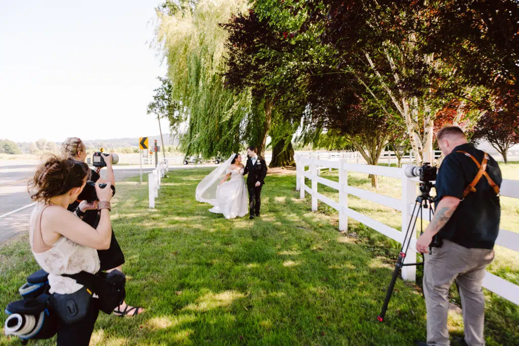 Snohomish Wedding Photographer GSquared Weddings A bride and groom walk along a grassy path near a white fence with two photographers capturing the moment Trees provide shade in the background and the sky is clear and sunny Serving Seattle Snohomish and Washington State