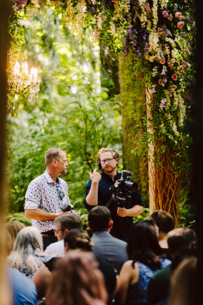 Snohomish Wedding Photographer GSquared Weddings Two men converse under a floral arch at a forest event. One holds a camera, indicating a focus on photography or filming. Guests are seated, all surrounded by lush greenery and hanging flowers, creating a vibrant, natural setting. Serving Seattle, Snohomish and Washington State