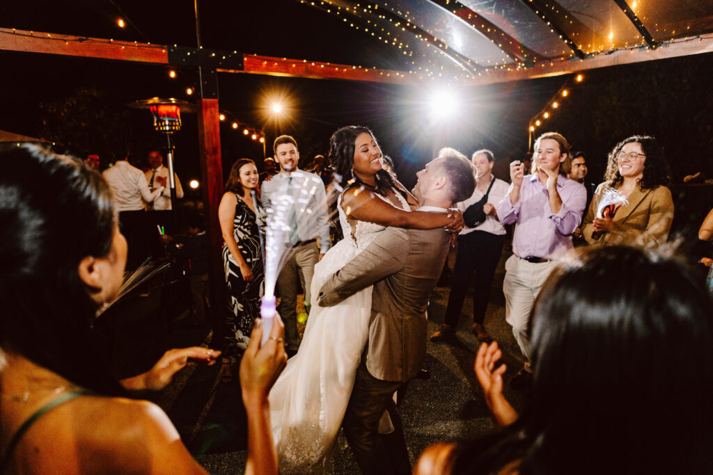 Snohomish Wedding Photographer GSquared Weddings A groom joyfully lifts the bride on a budget-friendly dance floor, surrounded by smiling guests at an outdoor evening celebration. String lights illuminate the scene, creating a festive atmosphere as they all cheer and capture the moment with their phones. Serving Seattle, Snohomish and Washington State