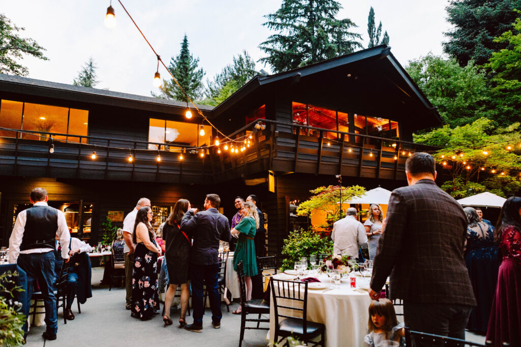 Snohomish Wedding Photographer GSquared Weddings A lively outdoor gathering at a house features guests mingling near tables with lights strung above. The backdrop is a large two-story building surrounded by trees. Everyone is dressed in formal attire, suggesting a celebratory event or party. Serving Seattle, Snohomish and Washington State
