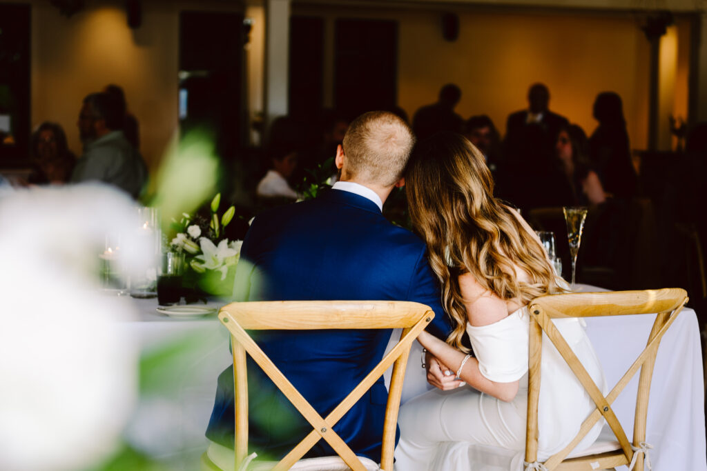 Snohomish Wedding Photographer GSquared Weddings A couple sits closely together on wooden chairs at a wedding reception The man wears a blue suit and the woman dazzles in an offshoulder white dress Theyre holding hands facing away from the camera with blurred guests and elegant decor in the background Serving Seattle Snohomish and Washington State Snohomish Wedding Photographer GSquared Weddings A couple sits closely together on wooden chairs at a wedding reception. The man wears a blue suit, and the woman dazzles in an off-shoulder white dress. Theyre holding hands, facing away from the camera, with blurred guests and elegant decor in the background. Serving Seattle, Snohomish and Washington State