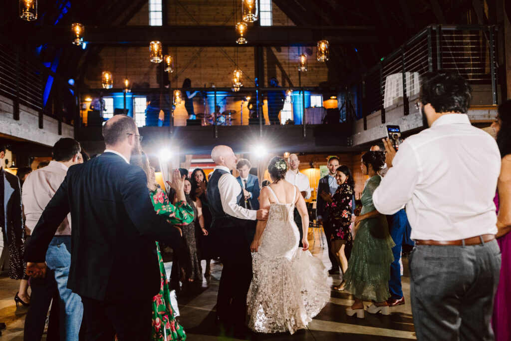 Snohomish Wedding Photographer GSquared Weddings A newlywed couple dances in the center of a rustic, warmly lit reception hall, surrounded by delighted guests. Some clap along while others capture photos, all beneath high ceilings adorned with hanging lights and an inviting open upper level. Serving Seattle, Snohomish and Washington State